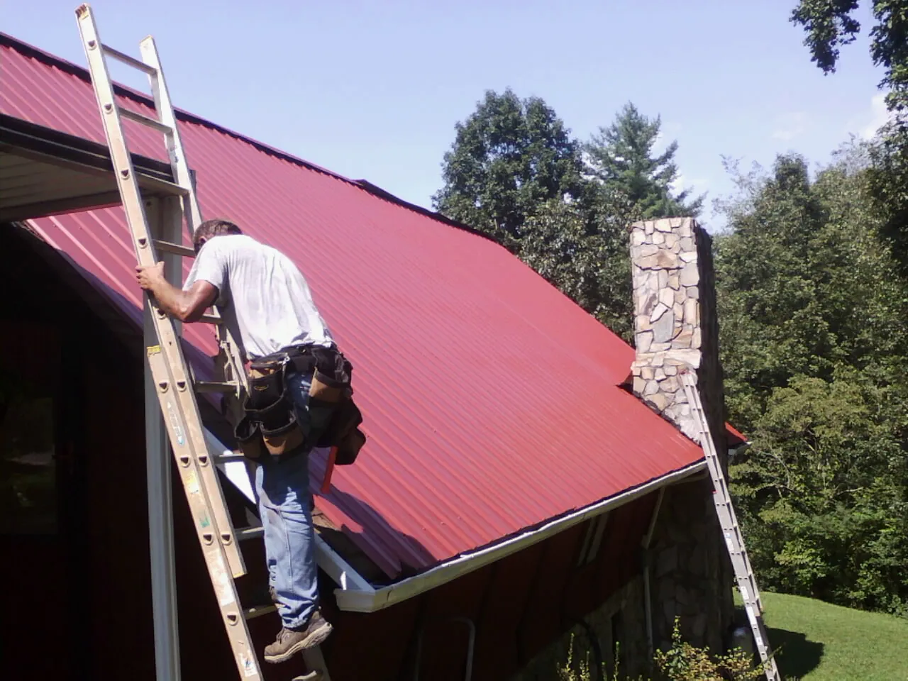 Roofer on a ladder beside a red metal roof and stone chimney — Soffit & Fascia Repair by Roofing Brothers in Huntsburg